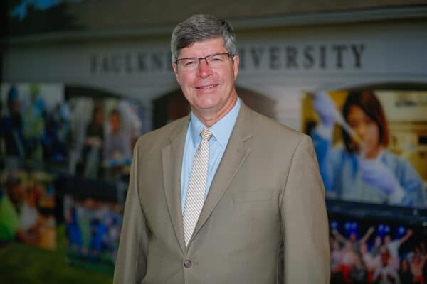 Faulkner University President Mitch Henry standing indoors, wearing a tan suit and striped tie, smiling in front of a university backdrop featuring campus and student images.