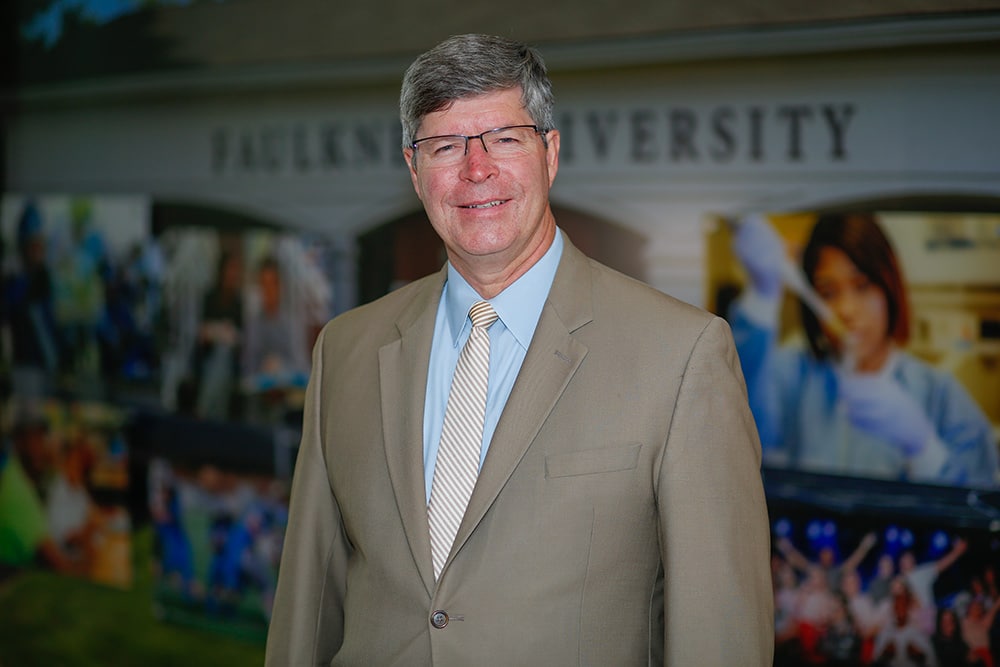 Faulkner University President Mitch Henry standing indoors, wearing a tan suit and striped tie, smiling in front of a university backdrop featuring campus and student images.