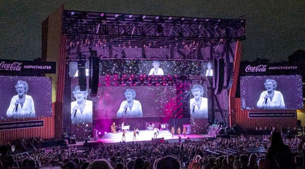 Rod Stewart performing on stage at the Coca-Cola Amphitheater in Birmingham, AL, with large video screens showing close-ups of him singing. The venue is packed with a large audience, and the stage is lit with bright pink and white lights. The amphitheater signage reads "Coca-Cola Amphitheater – Birmingham’s Home for Live Music"